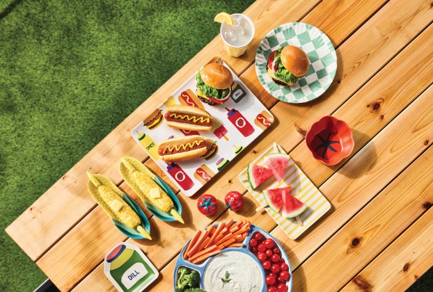 A picnic table set with burgers, hot dogs, corn holders, dip and watermelon slices on colorful, graphic tableware.