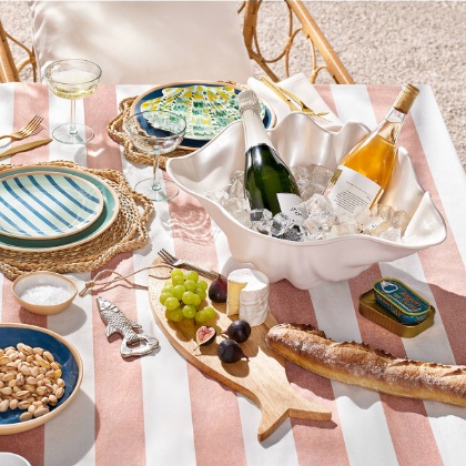 A summer dining setup featuring champagne in a shell-shaped bowl, rattan placemats, striped plates and a fish-shaped cutting board with cheese and fruit.