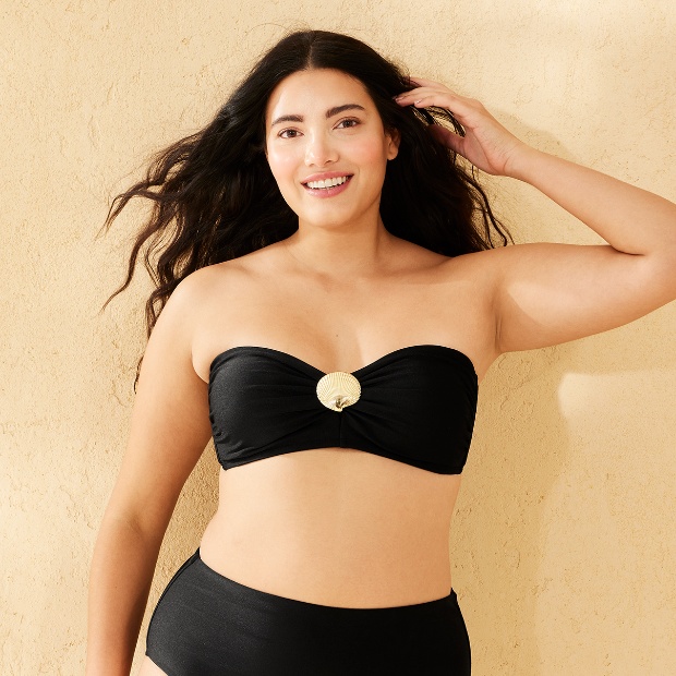 A person models a strapless black bandeau bikini top with a metallic gold shell accent at the center, paired with high-waisted bottoms.