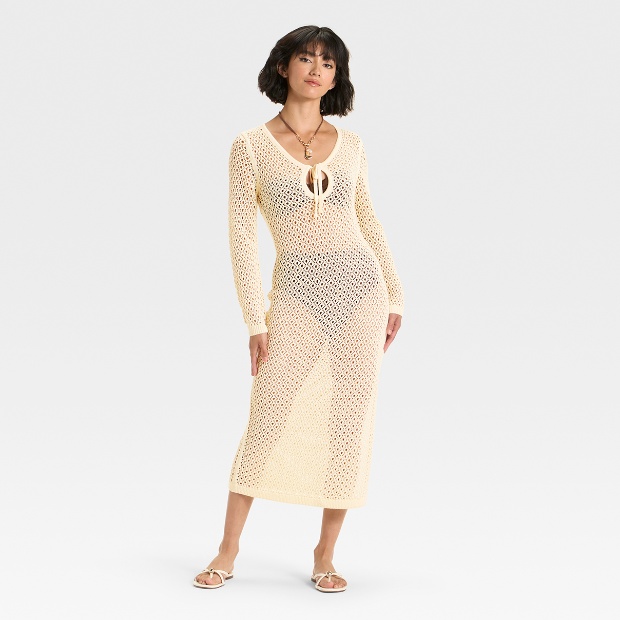 A person wears a long, ivory crochet-style maxi dress over a swimsuit with a tie-front neckline and long sleeves.