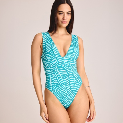 A person models a teal one-piece swimsuit with a deep V neckline and a jade palm print, standing against a light neutral backdrop.